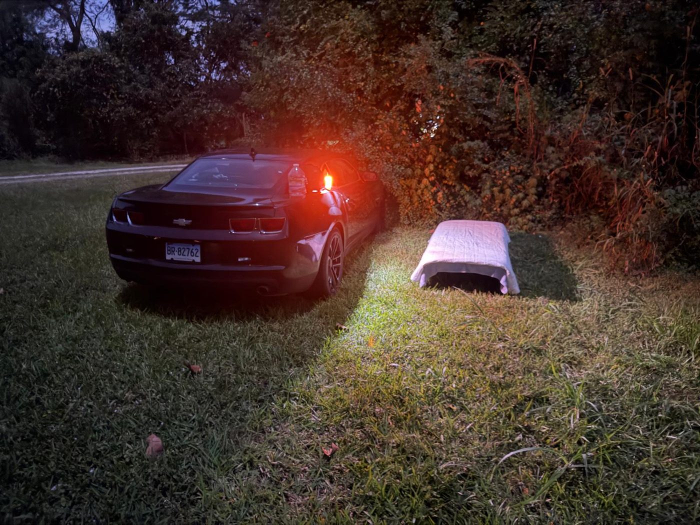 A cot set up on grass beside a car at night, lit by the car's headlights.